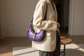 Purple printed leather crescent shoulder bag worn over the shoulder with a neutral tailored outfit