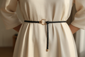 Slim black braided belt with gold-tone O-ring closure worn at the waist over an ivory dress